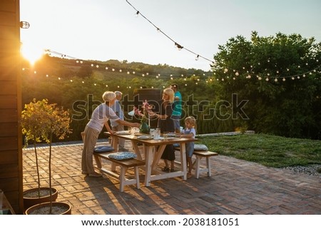 Whole family participates in the organization of dinner. Three female members setting dining table outdoors and male members barbecuing near them