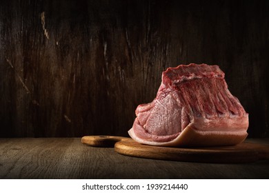 a whole freshly cut piece of raw pork loin lies on a cutting board on a brown wooden background in semidarkness. side view. artistic moody photo in simple rustic style with copy space