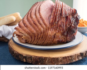 A Whole Glazed And Freshly Baked Spiral Cut Ham Sits On A Platter, The Slices Falling Away From The Bone, Accompanied By A Basket Of Breadsticks And A Bowl Of Mashed Yams In The Background