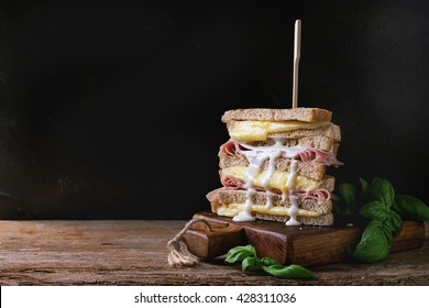 Whole Grain Grilled Sandwich Bread With Melting Hot Cheese, Ham, Basil And Pouring White Sauce On Wooden Chopping Board Over Dark Background. With Copy Space