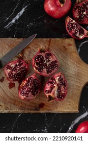 Whole And Halved Pomegranates On A Wooden Cutting Board With A Knife And Spilled Juice Placed On A Dark Background. Vertical Shot. Top Table View, Flat Lay.