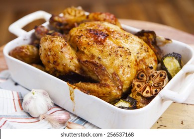Whole roasted chicken with garlic, potatoes and vegetables in a white ceramic roasting pan