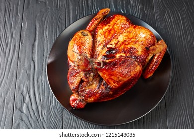 whole roasted chicken with golden brown crispy skin on a black plate on a wooden table, view from above