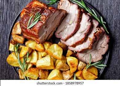 Whole roasted pork loin served with baked potato wedges, rosemary  on a black plate on a dark wooden background, top view, close up