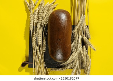 A whole rye bread loaf placed on a wooden board surrounded by wheat stalks on a vibrant yellow background, highlighting natural ingredients and rustic aesthetics - Powered by Shutterstock