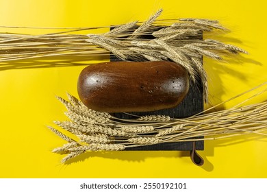 A whole rye bread loaf placed on a wooden board surrounded by wheat stalks on a vibrant yellow background, highlighting natural ingredients and rustic aesthetics - Powered by Shutterstock