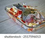 Whole Seasoned Sea ​​Bream Fish Baked with Fresh Vegetables Including Tomatoes, Potatoes, and Onions in a Glass Tray on Wooden Countertop, Ready for Roasting in a Home Kitchen