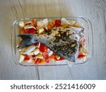 Whole Seasoned Sea ​​Bream Fish Baked with Fresh Vegetables Including Tomatoes, Potatoes, and Onions in a Glass Tray on Wooden Countertop, Ready for Roasting in a Home Kitchen