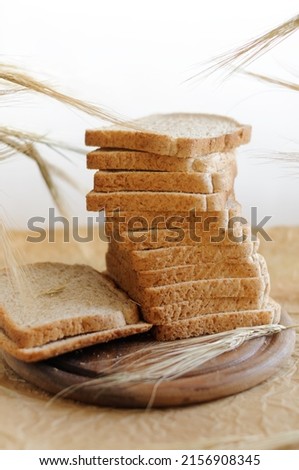 Whole wheat toast slices stacked with ears of rye on a round wooden board with parchment and a light gray background. Selective focus at shallow depth of field