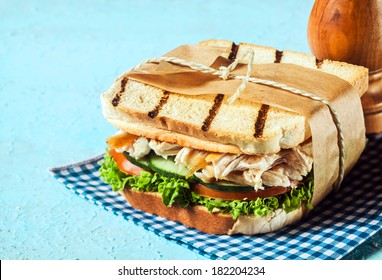 Wholesome Country Toasted Sandwich In A Wrapper With Shredded Chicken, Frilly Lettuce, Cucumber And Tomato Served On A Blue And White Checked Napkin On A Blue Background