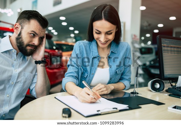 Why are we doing this? Unhappy\
and anxious bearded man covering his face with hand looks like his\
wife with a smile signs a contract to buy a car in\
dealership.
