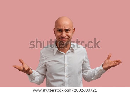 Why you do this. Horizontal shot of bald guy with bristle. With puzzled expression, spreads hands with confused look, frowns face, feels confused. Dressed in white shirt, isolated on pink background
