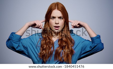Wicked red hair girl looking at camera in studio. Closeup angry woman putting fingers in ears on gray background. Portrait of agitated female person shaking head indoors.