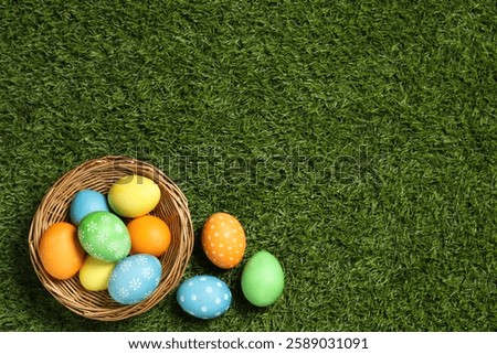 Wicker basket with decorated Easter eggs on green grass, top view. Space for text