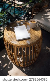 Wicker coffee table features a blank notebook and a cup of coffee, set amidst green plants and a swimming pool visible in the background