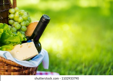 Wicker Picnic Basket With Cheese And Wine On Red Checkered Table Cloth On Green Grass Outside In Summer Park, No People