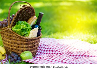 Wicker Picnic Basket With Cheese And Wine On Red Checkered Table Cloth On Green Grass Outside In Summer Park, No People