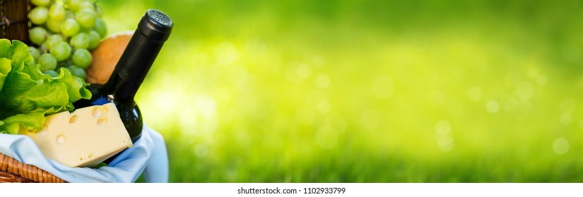 Wicker Picnic Basket With Cheese And Wine On Red Checkered Table Cloth On Green Grass Outside In Summer Park, No People