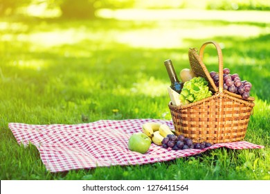 Wicker Picnic Basket With Cheese And Wine On Red Checkered Table Cloth On Green Grass Outside In Summer Park, No People