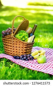 Wicker Picnic Basket With Cheese And Wine On Red Checkered Table Cloth On Green Grass Outside In Summer Park, No People