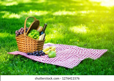 Wicker Picnic Basket With Cheese And Wine On Red Checkered Table Cloth On Green Grass Outside In Summer Park, No People