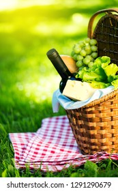 Wicker Picnic Basket With Cheese And Wine On Red Checkered Table Cloth On Green Grass Outside In Summer Park, No People