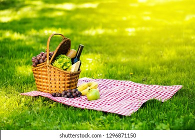 Wicker Picnic Basket With Cheese And Wine On Red Checkered Table Cloth On Green Grass Outside In Summer Park, No People