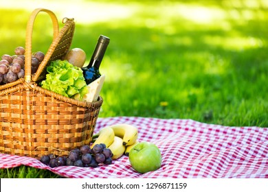 Wicker Picnic Basket With Cheese And Wine On Red Checkered Table Cloth On Green Grass Outside In Summer Park, No People