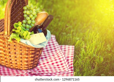 Wicker Picnic Basket With Cheese And Wine On Red Checkered Table Cloth On Green Grass Outside In Summer Park, No People