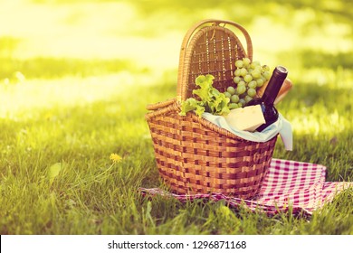 Wicker Picnic Basket With Cheese And Wine On Red Checkered Table Cloth On Green Grass Outside In Summer Park, No People