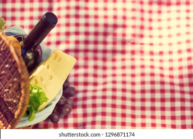 Wicker Picnic Basket With Cheese And Wine On Red Checkered Table Cloth Outside, Top View With Copy Space, No People