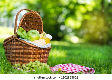 Wicker Picnic Basket With Healthy Food On Red Checkered Table Cloth On Green Grass Outside In Summer Park, No People