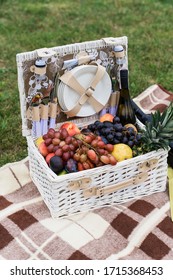 Wicker Picnic Basket With Plates, Fruits And Wine On Red Checkered Blanket On Green Grass Outside In Summer Park, No People