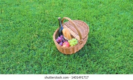 
Wicker Picnic Basket With White And Black Grapes And Wine On Green Grass Outside In Summer Park, No People