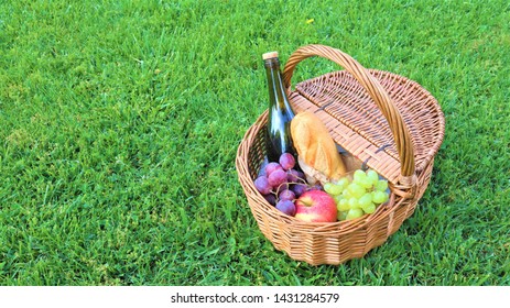 
Wicker Picnic Basket With White And Black Grapes And Wine On Green Grass Outside In Summer Park, No People