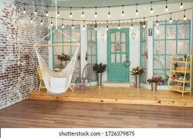 Wicker Swing-hammock On The Background Of The Summer Porch Of The House. Swing And Summer Terrace With Light Bulbs Retro Garlands. Beautiful Porch With Spring Decorations In A Photo Studio