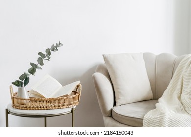 Wicker tray and open book close to eucalyptus branch in ceramic vase on side table. Bohemian design of living room with beige soft chair with cushion and white plaid - Shutterstock ID 2184160043