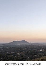 Wide aerial angle view of a small mountain ridge over a lush green village during a calm sunrise or sunset. Minimalist gradient sky with a solitary flying bird, perfect for tranquility, nature, and travel background concept.