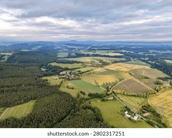 Wide aerial view showing patchwork green and golden fields, dense pine forest, scattered farmhouses, and curving hills around small rural village under soft cloudy dusk light with long shadows