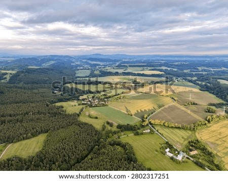 Wide aerial view showing patchwork green and golden fields, dense pine forest, scattered farmhouses, and curving hills around small rural village under soft cloudy dusk light with long shadows