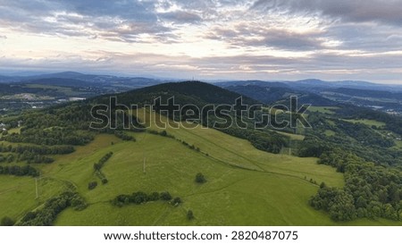Wide aerial view shows patchwork farmland, wooded hills and distant villages beneath soft clouded sky, with central hill, transmission tower and powerlines adding depth and scale, with landscape