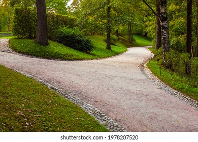 A wide alley in the park is divided into two paths diverging in different directions. The paths are lined with stone gutters.