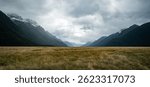 A wide alpine like valley with golden grasses stretches between steep, forested mountains shrouded in low clouds, with snow-capped peaks and a moody sky hinting at changing weather.