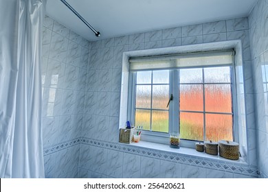 Wide angle bathroom interior, inc edge of shower curtain and curtain rail, tiled walls, frosted glass window with distorted exterior, and various items on the windowsill [landscape format].