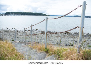 Wide angle close up on concrete stairs with a chain link railing, lined by tall grass, leading down to a rocky beach park, with a coastal cove in background - Powered by Shutterstock