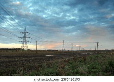 Wide angle of electrical power lines at dusk with a blue and pink sky. The lines lead away across a muddy field, to a vanishing point in the distance.