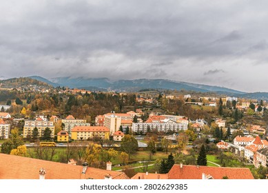 Wide angle elevated view of a Bohemian town in the Czech Republic during autumn. The scene shows a mix of red roofed family houses, apartment buildings and civic structures surrounded by green parks, trees and forested hillsides. A small river and road with cars and a yellow bus run through the town. In the background fog and low clouds cover the distant mountains, creating an atmospheric overcast mood with vibrant fall foliage colors.