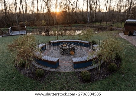 Wide angle elevated view shows a circular stone fire pit, concentric wooden benches, a pond, trimmed shrubs, frosted grass, and a path to a deck and hot tub at sunset.