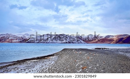 Wide Angle image of Pangong Lake in winters. Landscape with pink textured mountains and lake in front. A thin layer of snow on ground and hills. Cloudy weather