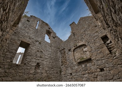 Wide angle low angle view looking up at the stone walls inside the roofless ruins of a medieval building. Historic limestone masonry of Penmon Priory with open window frames against a blue sky with wispy clouds on the Isle of Anglesey, North Wales.